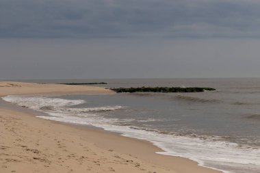 Jersey sahilinin güzel bir görüntüsü. Dalgaların beyaz köpüğü kumsalın güzel kumlarına çarpıyor. Siyah taşlı küçük bir iskele görülebilir. Mavi gökyüzünde beyaz bulutlar var..