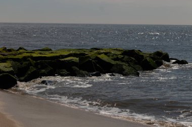 Bu, suya uzanan bir iskelenin resmi. Dalgalar etrafa köpük saçıyor. Körfezden çıkan büyük siyah kayaların üzerinde yeşil yosun yetişiyor..