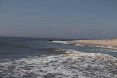 Jersey sahilinin güzel bir görüntüsü. Dalgaların beyaz köpüğü kumsalın güzel kumlarına çarpıyor. Siyah taşlı küçük bir iskele görülebilir. Mavi gökyüzünde beyaz bulutlar var..