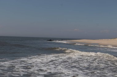 Jersey sahilinin güzel bir görüntüsü. Dalgaların beyaz köpüğü kumsalın güzel kumlarına çarpıyor. Siyah taşlı küçük bir iskele görülebilir. Mavi gökyüzünde beyaz bulutlar var..