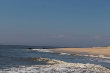 Jersey sahilinin güzel bir görüntüsü. Dalgaların beyaz köpüğü kumsalın güzel kumlarına çarpıyor. Siyah taşlı küçük bir iskele görülebilir. Mavi gökyüzünde beyaz bulutlar var..