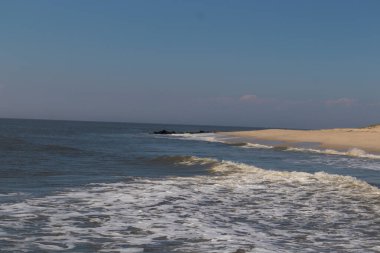 Jersey sahilinin güzel bir görüntüsü. Dalgaların beyaz köpüğü kumsalın güzel kumlarına çarpıyor. Siyah taşlı küçük bir iskele görülebilir. Mavi gökyüzünde beyaz bulutlar var..