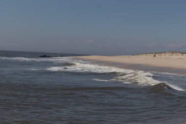 Jersey sahilinin güzel bir görüntüsü. Dalgaların beyaz köpüğü kumsalın güzel kumlarına çarpıyor. Siyah taşlı küçük bir iskele görülebilir. Mavi gökyüzünde beyaz bulutlar var..