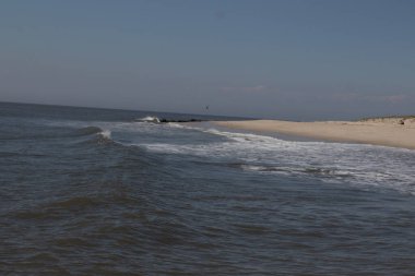 Jersey sahilinin güzel bir görüntüsü. Dalgaların beyaz köpüğü kumsalın güzel kumlarına çarpıyor. Siyah taşlı küçük bir iskele görülebilir. Mavi gökyüzünde beyaz bulutlar var..