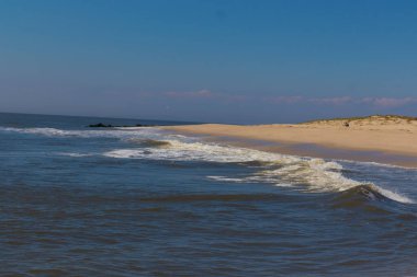 Jersey sahilinin güzel bir görüntüsü. Dalgaların beyaz köpüğü kumsalın güzel kumlarına çarpıyor. Siyah taşlı küçük bir iskele görülebilir. Mavi gökyüzünde beyaz bulutlar var..