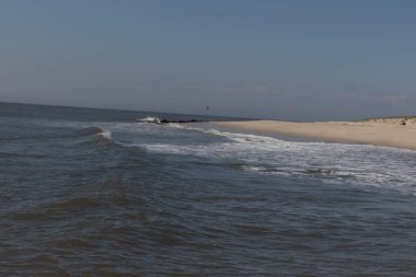Jersey sahilinin güzel bir görüntüsü. Dalgaların beyaz köpüğü kumsalın güzel kumlarına çarpıyor. Siyah taşlı küçük bir iskele görülebilir. Mavi gökyüzünde beyaz bulutlar var..