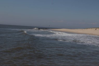 Jersey sahilinin güzel bir görüntüsü. Dalgaların beyaz köpüğü kumsalın güzel kumlarına çarpıyor. Siyah taşlı küçük bir iskele görülebilir. Mavi gökyüzünde beyaz bulutlar var..