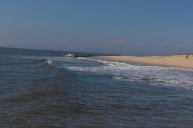 Jersey sahilinin güzel bir görüntüsü. Dalgaların beyaz köpüğü kumsalın güzel kumlarına çarpıyor. Siyah taşlı küçük bir iskele görülebilir. Mavi gökyüzünde beyaz bulutlar var..