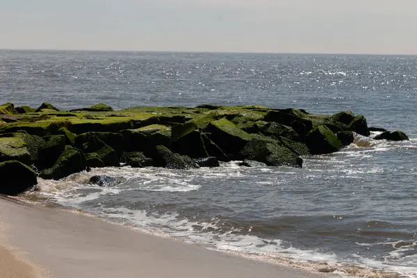 Bu, suya uzanan bir iskelenin resmi. Dalgalar etrafa köpük saçıyor. Körfezden çıkan büyük siyah kayaların üzerinde yeşil yosun yetişiyor..