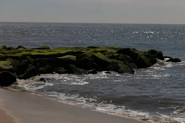 Bu, suya uzanan bir iskelenin resmi. Dalgalar etrafa köpük saçıyor. Körfezden çıkan büyük siyah kayaların üzerinde yeşil yosun yetişiyor..