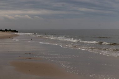 Bu plajın güzel bir gününün görüntüsü. Dalgaların yumuşak akıntıları kıyıya vurmaya geliyor, her geçişte kumu dışarı taşıyor. Bulutlar gökyüzüne yayıldı..