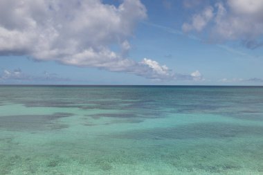 Sakin, turkuaz bir okyanusun soluk bir ufuk çizgisi olan geniş bir manzarası. Su berrak, aşağıdaki sualtı manzarasını ortaya çıkarıyor. Yukarıda, koyu mavi bir gökyüzü birkaç büyük, beyaz ve gri bulutla benekli..