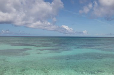 Sakin, turkuaz bir okyanusun soluk bir ufuk çizgisi olan geniş bir manzarası. Su berrak, aşağıdaki sualtı manzarasını ortaya çıkarıyor. Yukarıda, koyu mavi bir gökyüzü birkaç büyük, beyaz ve gri bulutla benekli..