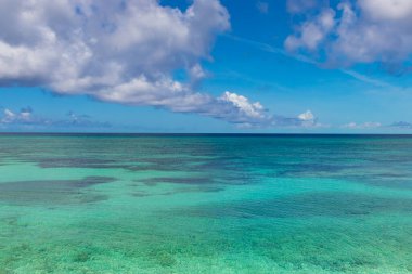 Sakin, turkuaz bir okyanusun soluk bir ufuk çizgisi olan geniş bir manzarası. Su berrak, aşağıdaki sualtı manzarasını ortaya çıkarıyor. Yukarıda, koyu mavi bir gökyüzü birkaç büyük, beyaz ve gri bulutla benekli..