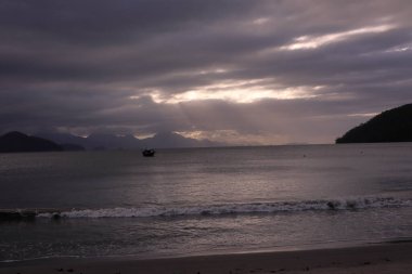 Brezilya, Ubatuba 'daki Itagua Sahili' nde huzurlu bir şafak, sakin okyanus suları sabah ışığını yansıtıyor. Sahne bulutlara, uzak dağlara ve tropikal bir kıyı şeridine sahipti. Huzur ve doğal güzellik için mükemmeldi..