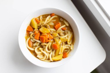 Japon körili udon, bir kase erişte ve havuçlu çorba. Kase beyazdır ve bir masaya oturur..