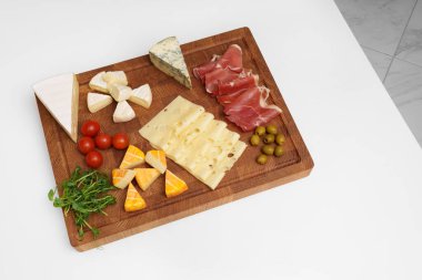 Peynir tahtası beyaz masada. Farklı peynir türleri, kırmızı biber, jamon, zeytin, domates, mikroyeşiller, bıçak, ekmek çubukları. Atıştırmalık Peynir parçaları..