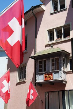Zürih 'te bir sokakta bulunan İsviçre bayrakları alçı pervazları ve altın bir aslan figürüyle süslenmiş dekoratif defne penceresi olan bir evin pembe cephesine doğru dalgalanıyor..