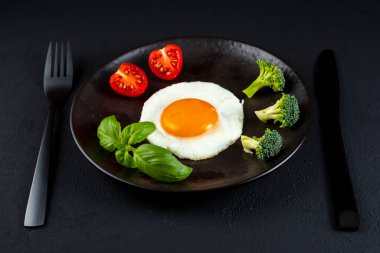 Fried soft-boiled egg on a black plate with fresh broccoli vegetables and tomatoes