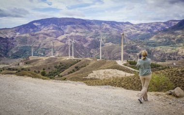 Sarışın genç bir kadın, rüzgar türbinli bir dağ manzarasının fotoğraflarını çekiyor.
