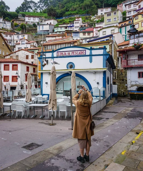 Asturias 'taki Cudillero kasabasında balık pazarının önündeki direğe tünemiş cep telefonuyla fotoğraf çeken kahverengi ceketli genç ve sarışın bir kadın.