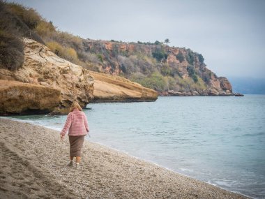 Pembe ceketli genç bir kadın. Sahilde yürüyor. Arka planda bir uçurum var.