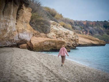 Pembe ceketli genç bir kadın. Sahilde yürüyor. Arka planda bir uçurum var.