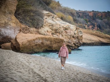 Pembe ceketli genç bir kadın. Sahilde yürüyor. Arka planda bir uçurum var.