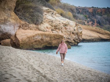 Pembe ceketli genç bir kadın. Sahilde yürüyor. Arka planda bir uçurum var.
