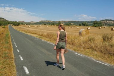 Arka planda ezilmiş saman balyalarıyla yolda yürüyen bir kadın.