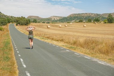 Arka planda ezilmiş saman balyalarıyla yolda yürüyen bir kadın.
