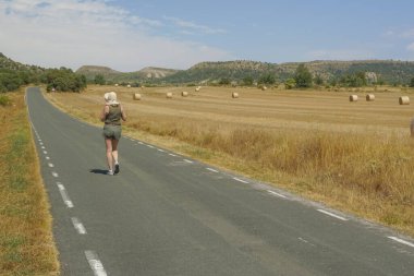 Arka planda ezilmiş saman balyalarıyla yolda yürüyen bir kadın.