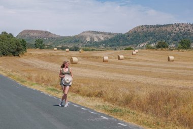 Arka planda ezilmiş saman balyalarıyla yolda yürüyen bir kadın.
