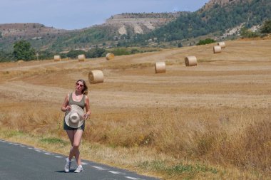 Arka planda ezilmiş saman balyalarıyla yolda yürüyen bir kadın.