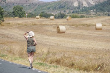 Arka planda ezilmiş saman balyalarıyla yolda yürüyen bir kadın.