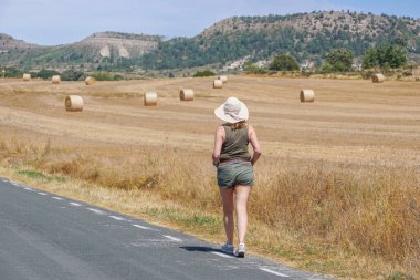 Arka planda ezilmiş saman balyalarıyla yolda yürüyen bir kadın.