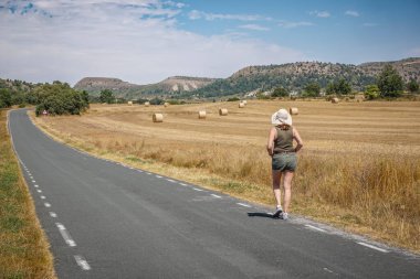 Arka planda ezilmiş saman balyalarıyla yolda yürüyen bir kadın.