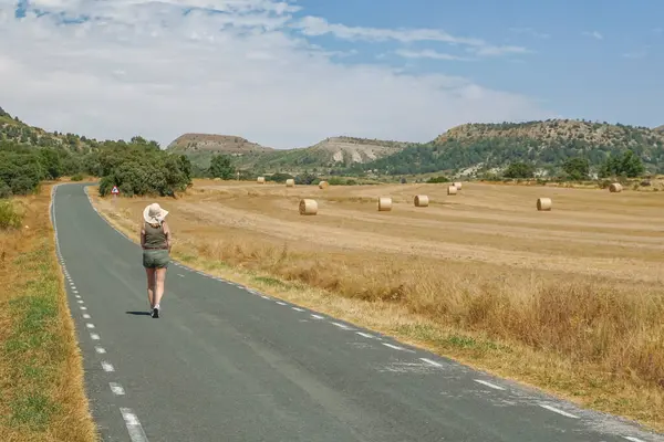 Arka planda ezilmiş saman balyalarıyla yolda yürüyen bir kadın.