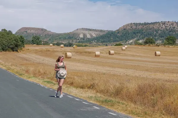 Arka planda ezilmiş saman balyalarıyla yolda yürüyen bir kadın.