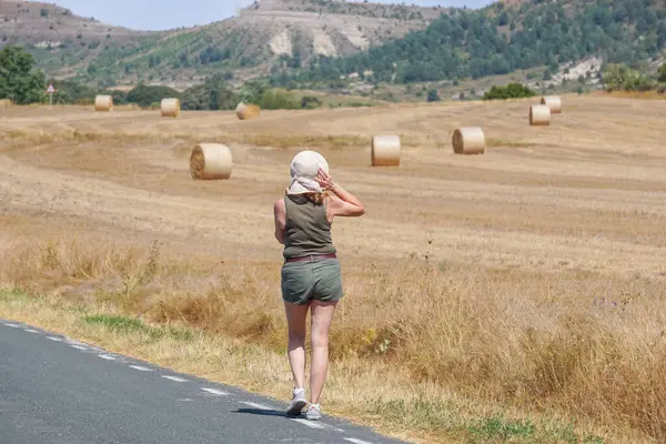 Arka planda ezilmiş saman balyalarıyla yolda yürüyen bir kadın.