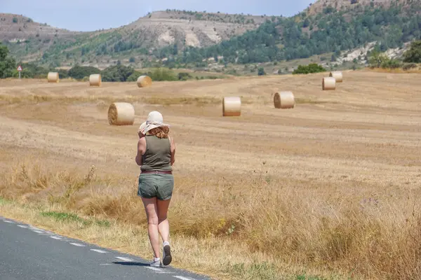 Arka planda ezilmiş saman balyalarıyla yolda yürüyen bir kadın.