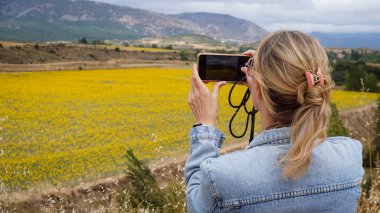 Mavi kot ceket giyen genç sarışın kadın cep telefonu kamerasıyla bir ayçiçeği tarlasının fotoğraflarını çekiyor.