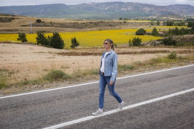 Ceket ve kot pantolon giyen genç sarışın kadın ıssız bir yolda yürüyor.