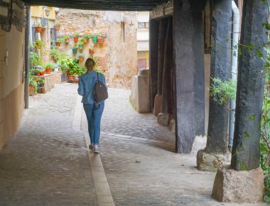 Ceket ve kot pantolon giyen genç sarışın kadın arka planda çiçek saksıları olan bir meydanda yürüyor.