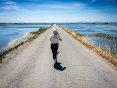 Şapkalı bir kadın ıssız bir yolda yürüyor, kenarlarında su var.
