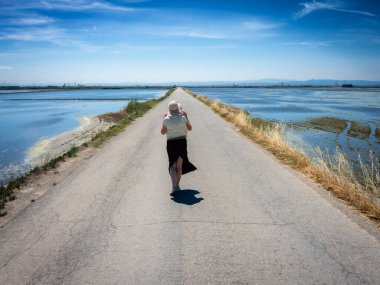 Şapkalı bir kadın ıssız bir yolda yürüyor, kenarlarında su var.