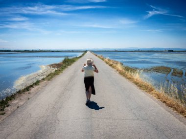 Şapkalı bir kadın ıssız bir yolda yürüyor, kenarlarında su var.