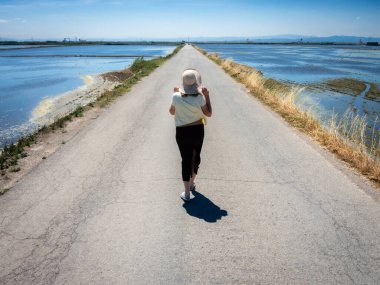 Şapkalı bir kadın ıssız bir yolda yürüyor, kenarlarında su var.