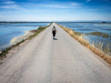 Şapkalı bir kadın ıssız bir yolda yürüyor, kenarlarında su var.