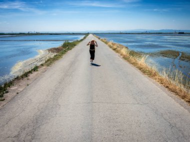 Şapkalı bir kadın ıssız bir yolda yürüyor, kenarlarında su var.
